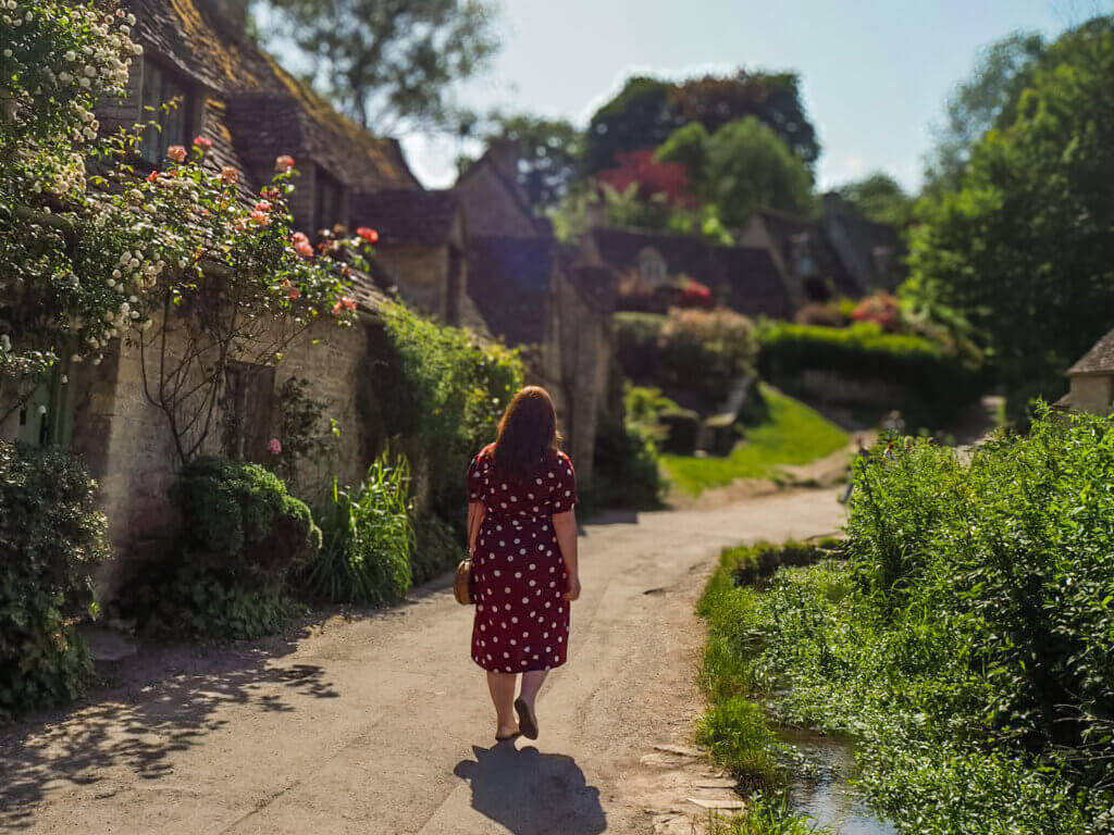 Nicola Lavin, an Irish travel blogger walks along a row of quaint stone cottages nestled in the lush greenery of the Cotswolds.