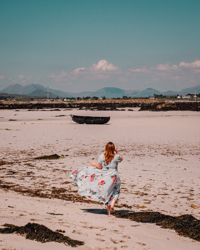 Nicola Lavin, an Irish travel blogger is wearing a blue floral dress and is walking along a white sandy beach in Connemara, Ireland.
