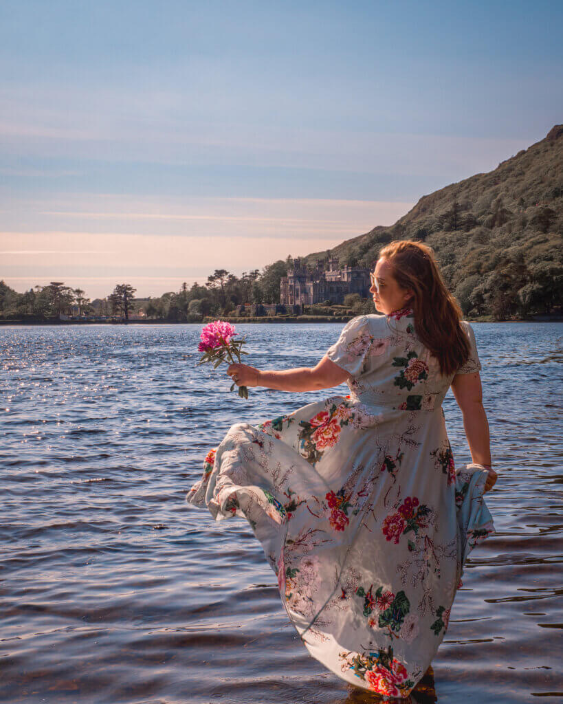 Nicola Lavin, an Irish travel blogger is wearing a blue floral dress. She is holding a pink flower and is standing barefoot in a lake in Connemara. Kylemore Abbey can be seen in the distance.