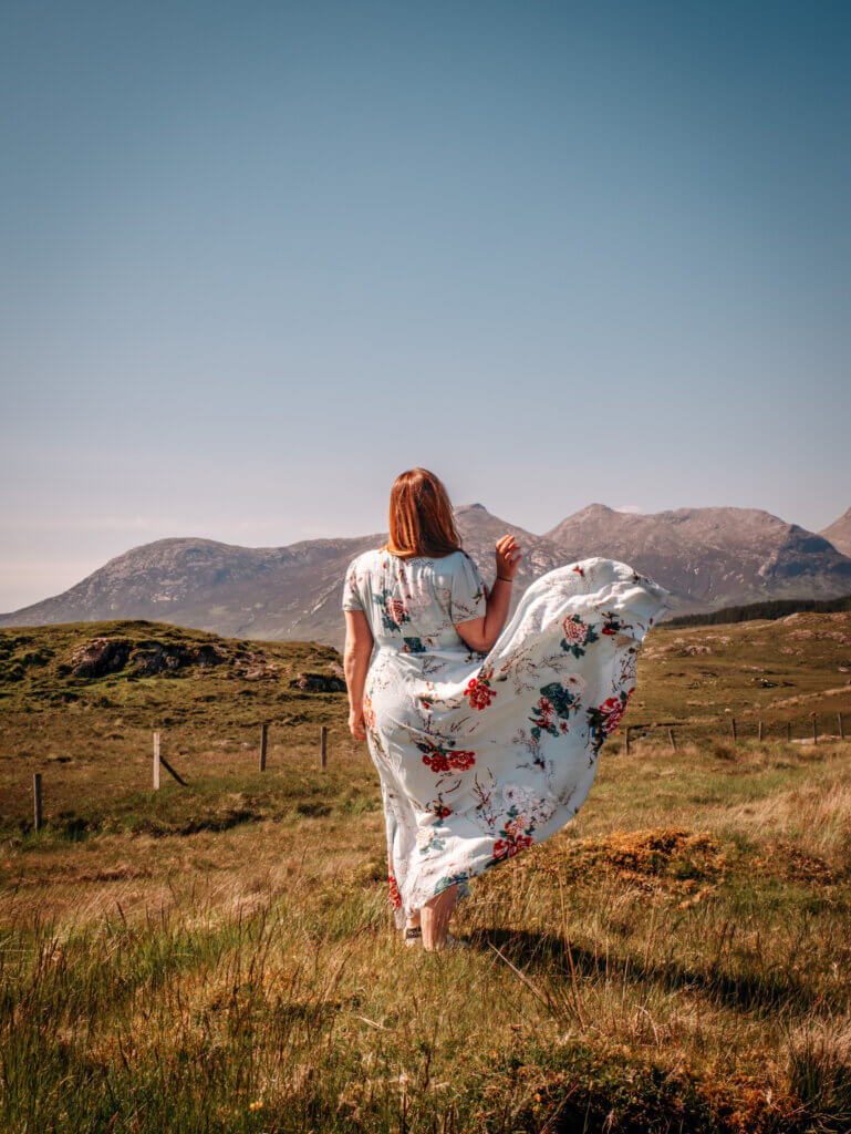 Nicola Lavin, an Irish travel blogger flicks her blue floral dress into the air. The background is the Twelve Ben mountain range in Connemara.