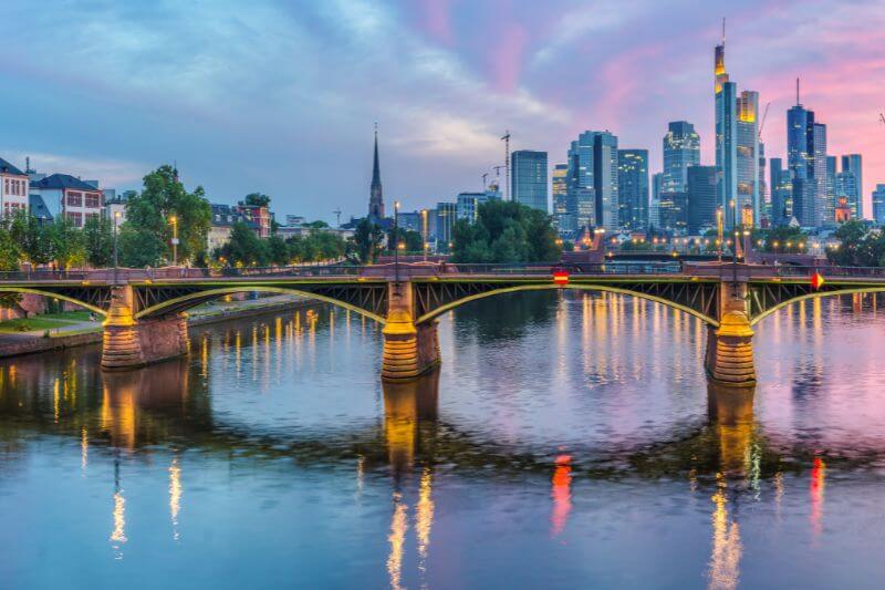 Bridge over the Main River in Frankfurt at sunset. Skyscraper buildings of the financial district are lit up in the background.