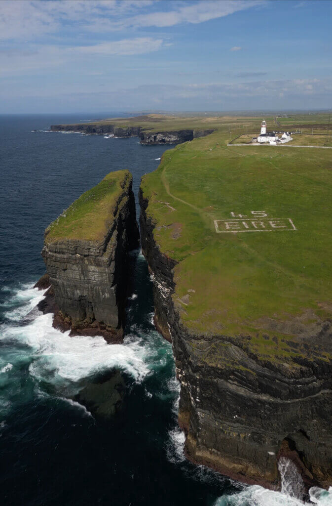 Aerial view of Loop Head Lighthouse and the Eire 45 sign. Waves crash around the sea stack at Loop Head peninsula.